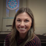 Smiling woman with long hair in a striped sweater, in a room with pews and a circular stained-glass window behind her.