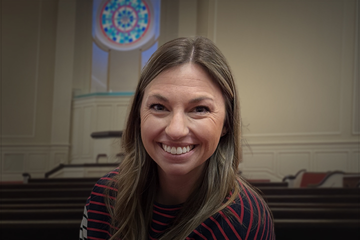 Smiling woman with long hair in a striped sweater, in a room with pews and a circular stained-glass window behind her.