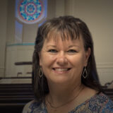Smiling woman with dark hair and diamond earrings in a softly lit room with a circular stained‑glass window behind her.