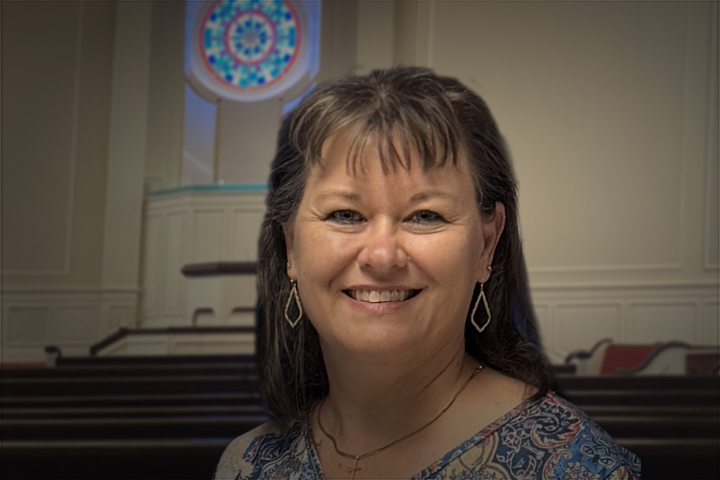 Smiling woman with dark hair and diamond earrings in a softly lit room with a circular stained‑glass window behind her.