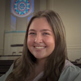 Smiling woman with long hair in a church, stained-glass window visible behind her.