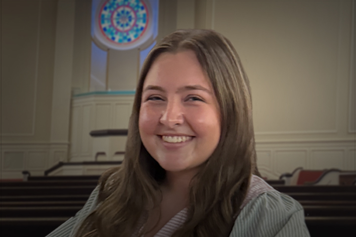 Smiling woman with long hair in a church, stained-glass window visible behind her.