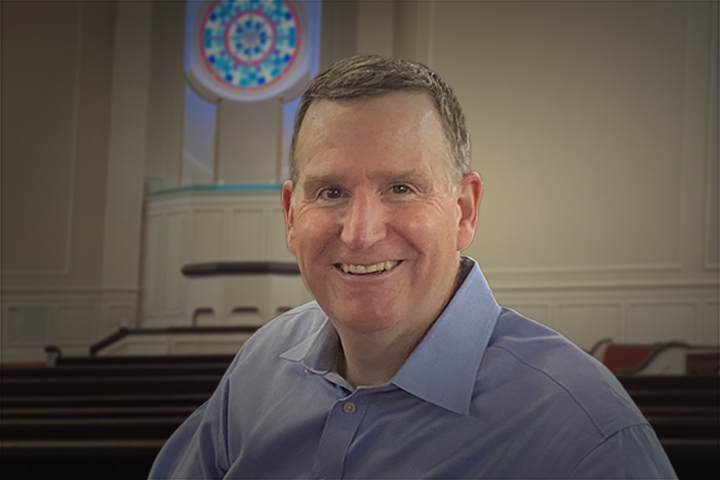 Smiling man in a light blue shirt posing in a church interior with wooden pews and a circular stained-glass window behind him