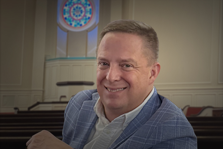 Smiling man in a light blue checked blazer and white shirt, seated in a church with a circular stained-glass window behind him.