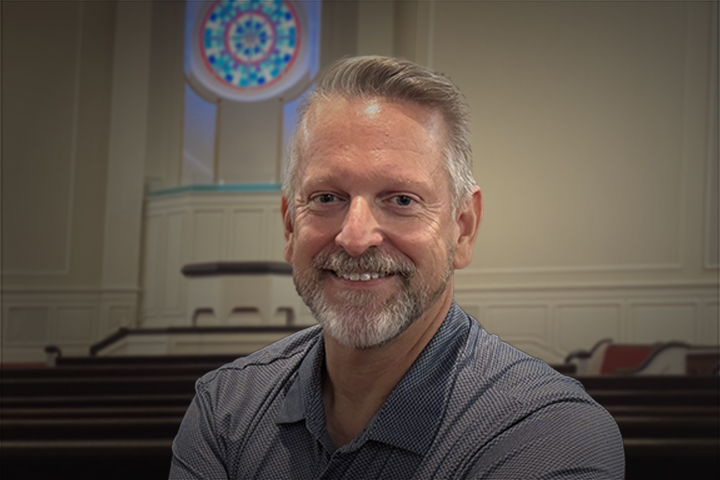 Smiling middle-aged man with gray hair and beard sits in a church sanctuary, with pews and a circular stained-glass window behind him.