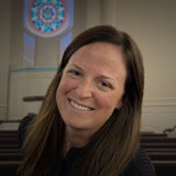 Smiling woman with long brown hair in a dark top, leaning to the side inside a church with pews and a circular stained-glass window behind her.
