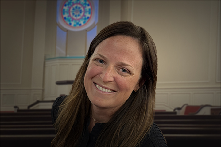 Smiling woman with long brown hair in a dark top, leaning to the side inside a church with pews and a circular stained-glass window behind her.
