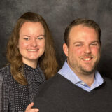 Two smiling adults posing together against a dark backdrop: woman on the left in a patterned blouse and man on the right in a light blue shirt and dark sweater.
