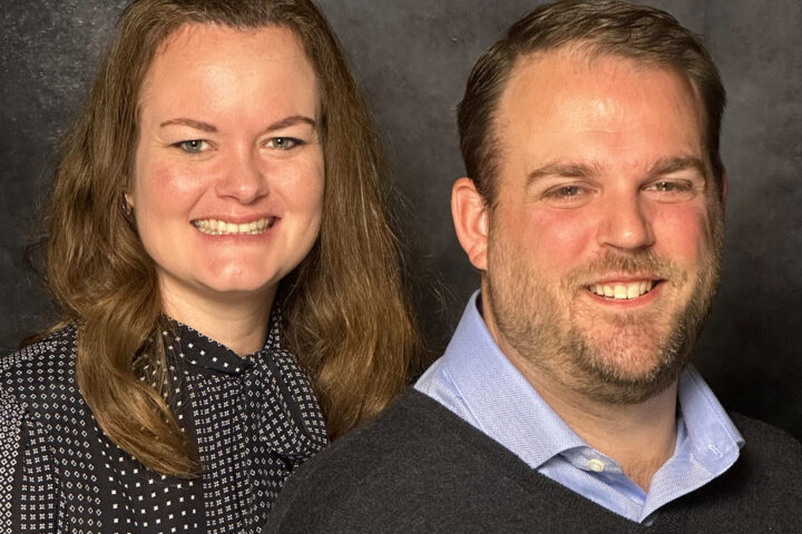 Two smiling adults posing together against a dark backdrop: woman on the left in a patterned blouse and man on the right in a light blue shirt and dark sweater.