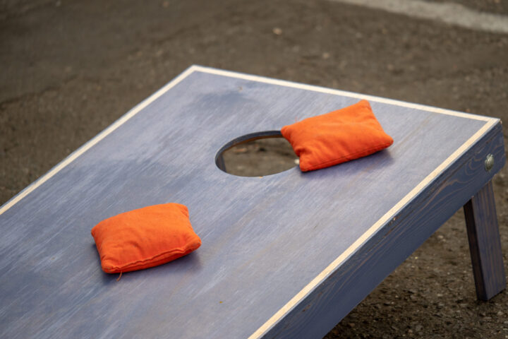 Blue wooden cornhole board with an orange bean bag resting near the hole outdoors.