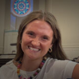 Smiling young woman in a white shirt wearing a colorful beaded necklace in a bright room with pews in the background, looking at the camera and leaning playfully to the side.