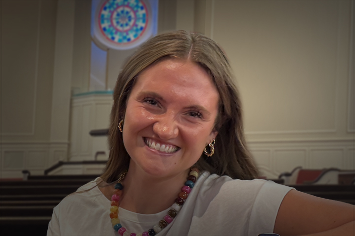 Smiling young woman in a white shirt wearing a colorful beaded necklace in a bright room with pews in the background, looking at the camera and leaning playfully to the side.
