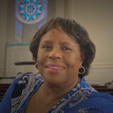 Smiling African American woman in a blue patterned blouse, inside a church with pews and a stained-glass circle in the background.