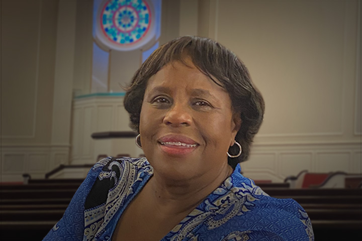 Smiling African American woman in a blue patterned blouse, inside a church with pews and a stained-glass circle in the background.