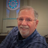 Smiling older man with gray hair and beard wearing glasses in a church sanctuary with stained-glass window behind him