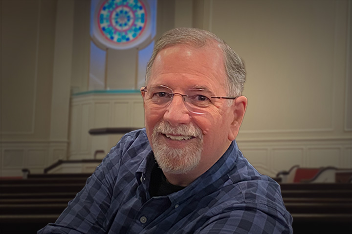 Smiling older man with gray hair and beard wearing glasses in a church sanctuary with stained-glass window behind him