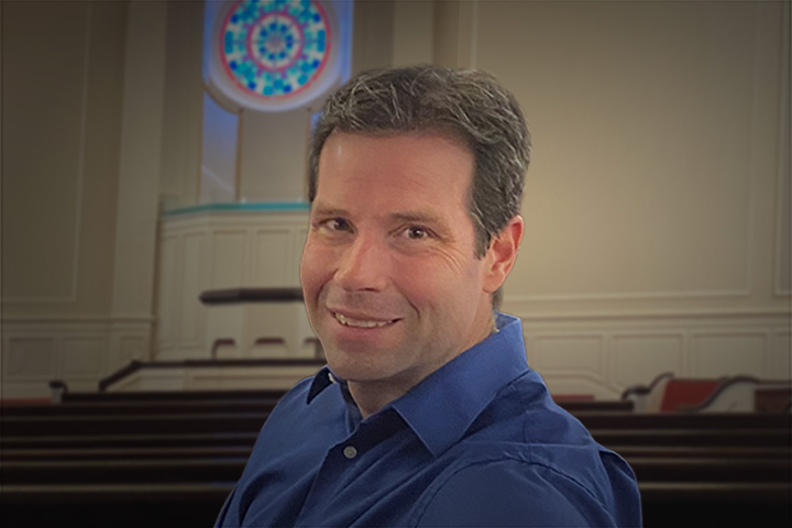 Smiling man in a blue shirt sits in a church sanctuary with pews and a circular stained-glass window behind him.