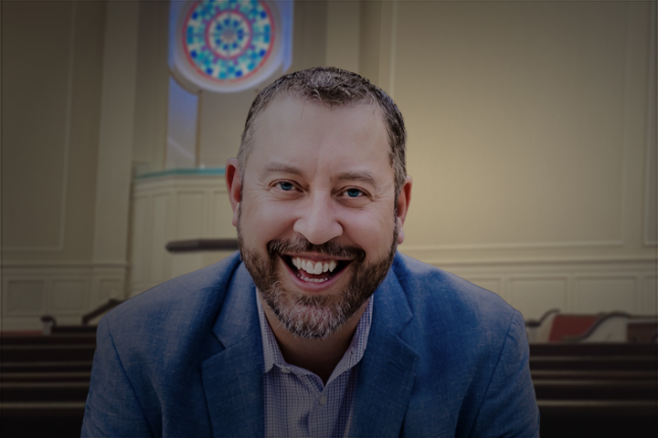 Smiling man in a blue blazer sits in a formal room with ornate wall paneling and a circular light fixture behind him.