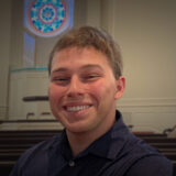 Young man smiling in a navy shirt, in a chapel with wooden pews and a circular stained‑glass window behind him.