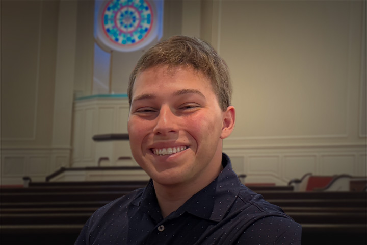 Young man smiling in a navy shirt, in a chapel with wooden pews and a circular stained‑glass window behind him.