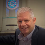 Smiling middle-aged man in a blazer seated at a pew inside a room with a stained glass window behind him