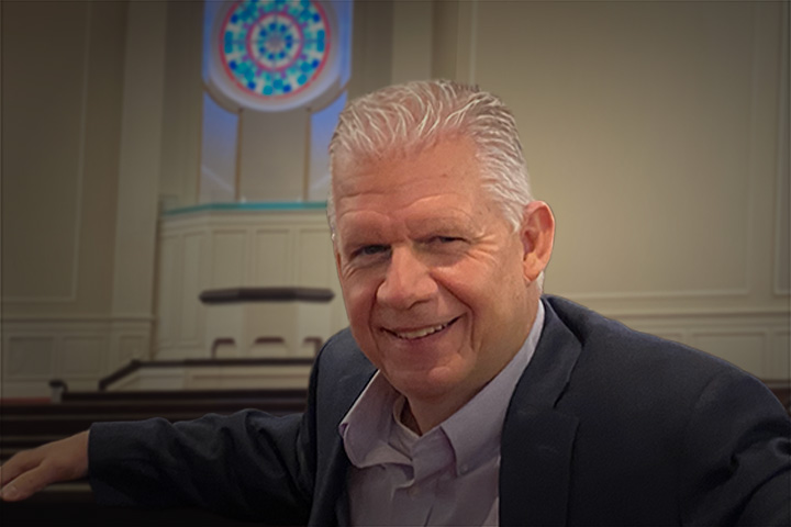 Smiling middle-aged man in a blazer seated at a pew inside a room with a stained glass window behind him