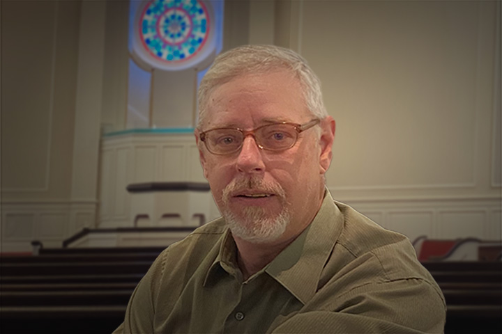 Middle-aged man with glasses and gray beard seated in a church sanctuary with a stained-glass window behind him.