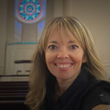 Smiling woman with blonde hair in a black shirt inside a church, a circular stained-glass window behind her.