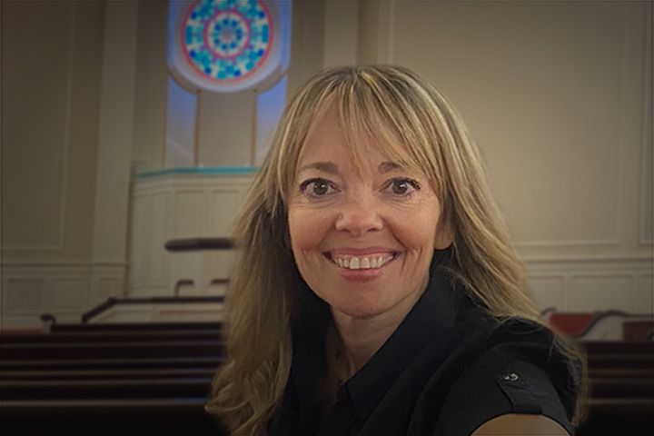 Smiling woman with blonde hair in a black shirt inside a church, a circular stained-glass window behind her.