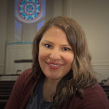Smiling woman with wavy hair wearing a maroon cardigan, indoors in a room with pews and a circular stained‑glass window behind her.