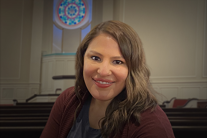 Smiling woman with wavy hair wearing a maroon cardigan, indoors in a room with pews and a circular stained‑glass window behind her.