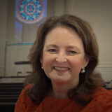 Portrait of a smiling woman with brown hair in a red blouse, seated in a formal room with pews and a circular stained-glass window in the background.