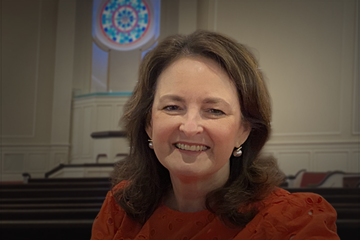 Portrait of a smiling woman with brown hair in a red blouse, seated in a formal room with pews and a circular stained-glass window in the background.