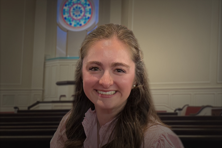 Smiling woman with long brown hair in a pink blouse, indoors near church pews and a circular stained-glass window behind her