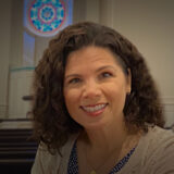 Portrait of a smiling woman with curly brown hair in a church sanctuary, pews visible in the background.