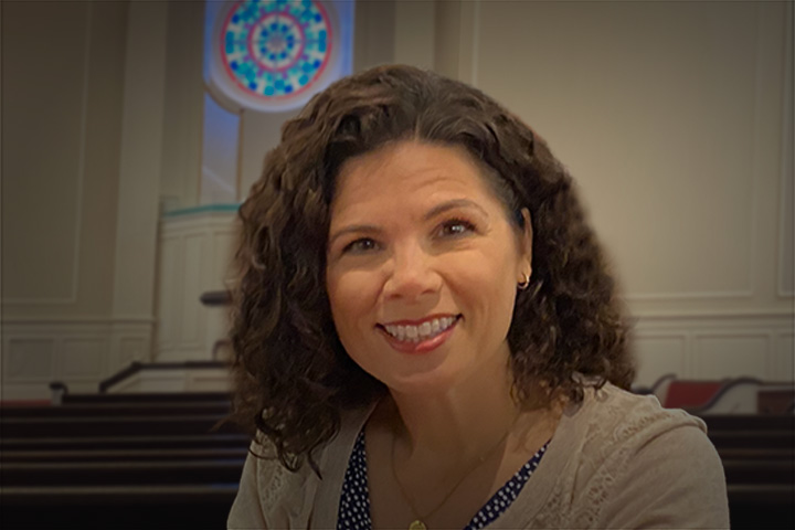 Portrait of a smiling woman with curly brown hair in a church sanctuary, pews visible in the background.