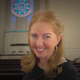 Smiling woman with light brown hair and gold hoop earrings in a church setting with pews in the background.