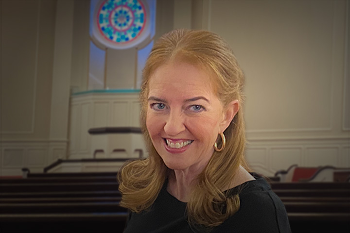 Smiling woman with light brown hair and gold hoop earrings in a church setting with pews in the background.