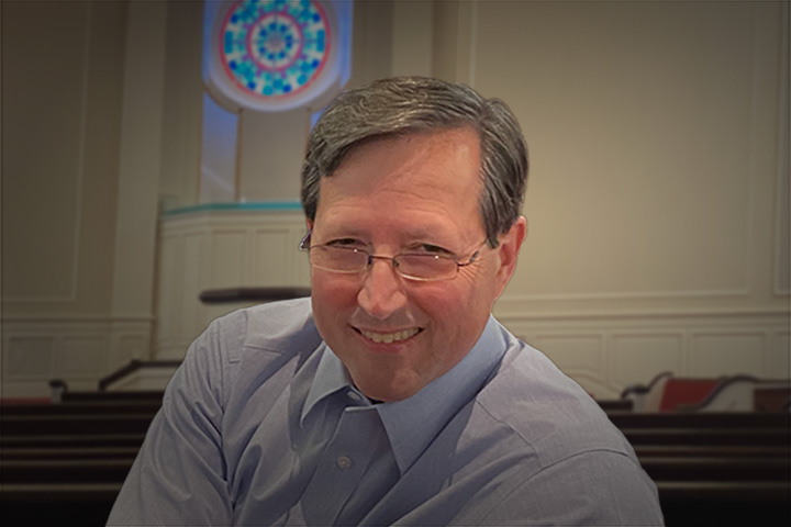 Smiling man with glasses in a light blue shirt, indoors near pews and a stained-glass window behind him.