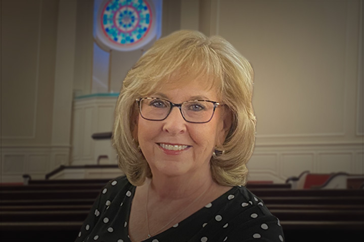 Portrait of a smiling woman with blonde hair and glasses in a church sanctuary background with pews and a stained-glass window behind her.