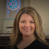 Smiling woman with shoulder-length hair in a church sanctuary with pews and a circular stained-glass window behind her in the background.