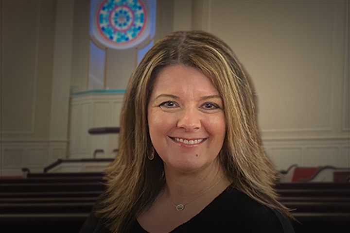 Smiling woman with shoulder-length hair in a church sanctuary with pews and a circular stained-glass window behind her in the background.