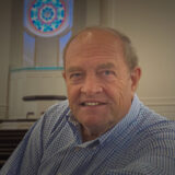 Smiling older man in a blue checkered shirt sits in a church pew with a stained-glass circle window behind him.